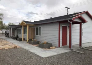 Two modern, single-story homes with grey siding, one featuring a yellow porch and the other with a red door, among gravel landscaping