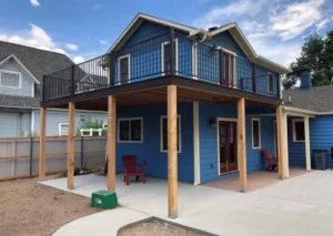 A two-story blue house with a balcony, supported by wooden posts, features a patio area and two red chairs outside