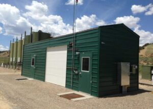 A long, green corrugated metal industrial building featuring a white roll-up garage door and two side entry doors