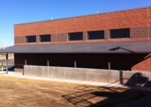 A modern brick building with a metal roof, featuring large windows and a grassy area in front, under a clear blue sky
