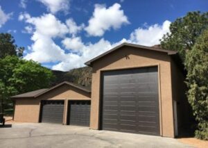 Two connected brown stucco garage buildings featuring three dark brown roll-up doors against a blue sky with white clouds