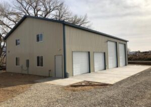 Large tan metal workshop building with four white roll-up garage doors of varying heights and a side entry door