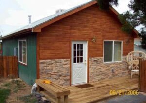 A cozy wood-sided cabin with a stone veneer base, a white cross-buck door, and a small wooden porch