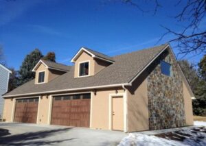 A large two-story tan garage and living space with a prominent stone accent wall, dormer windows, and two dark wood garage doors