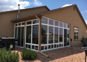 Exterior view of a tall, white-framed glass sunroom enclosure attached to a tan stucco home, set against a gravel backyard